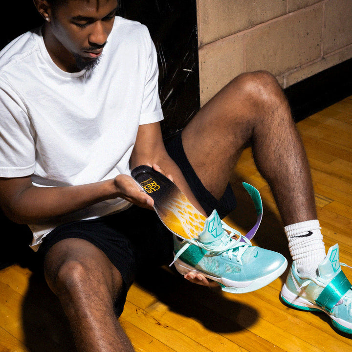 Person sitting on a wooden court floor wearing turquoise sneakers with white laces, placing CURREX BASKETBALLPRO into shoe #profile_high