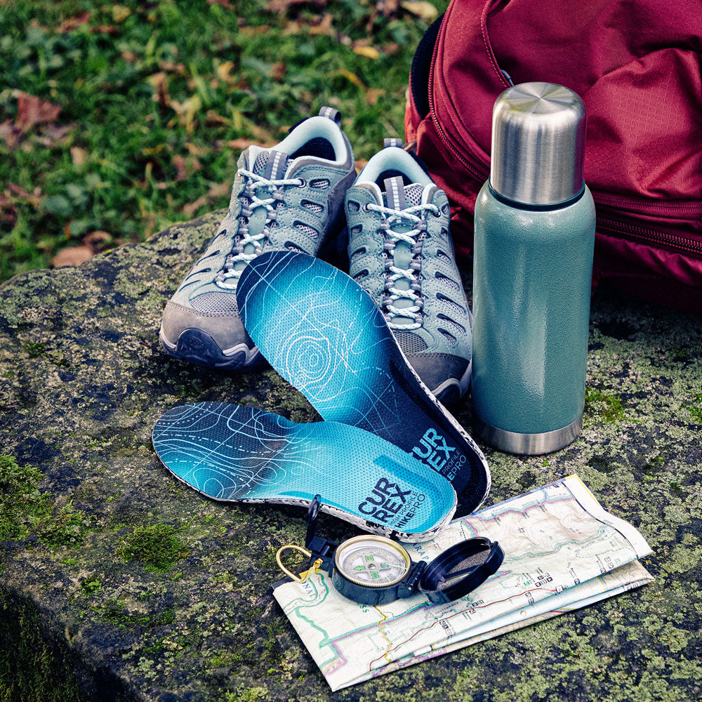CURREX HIKEPRO insoles next to hiking accessories: shoes, thermos, backpack, compass and map sitting on moss-covered rock #profile_high