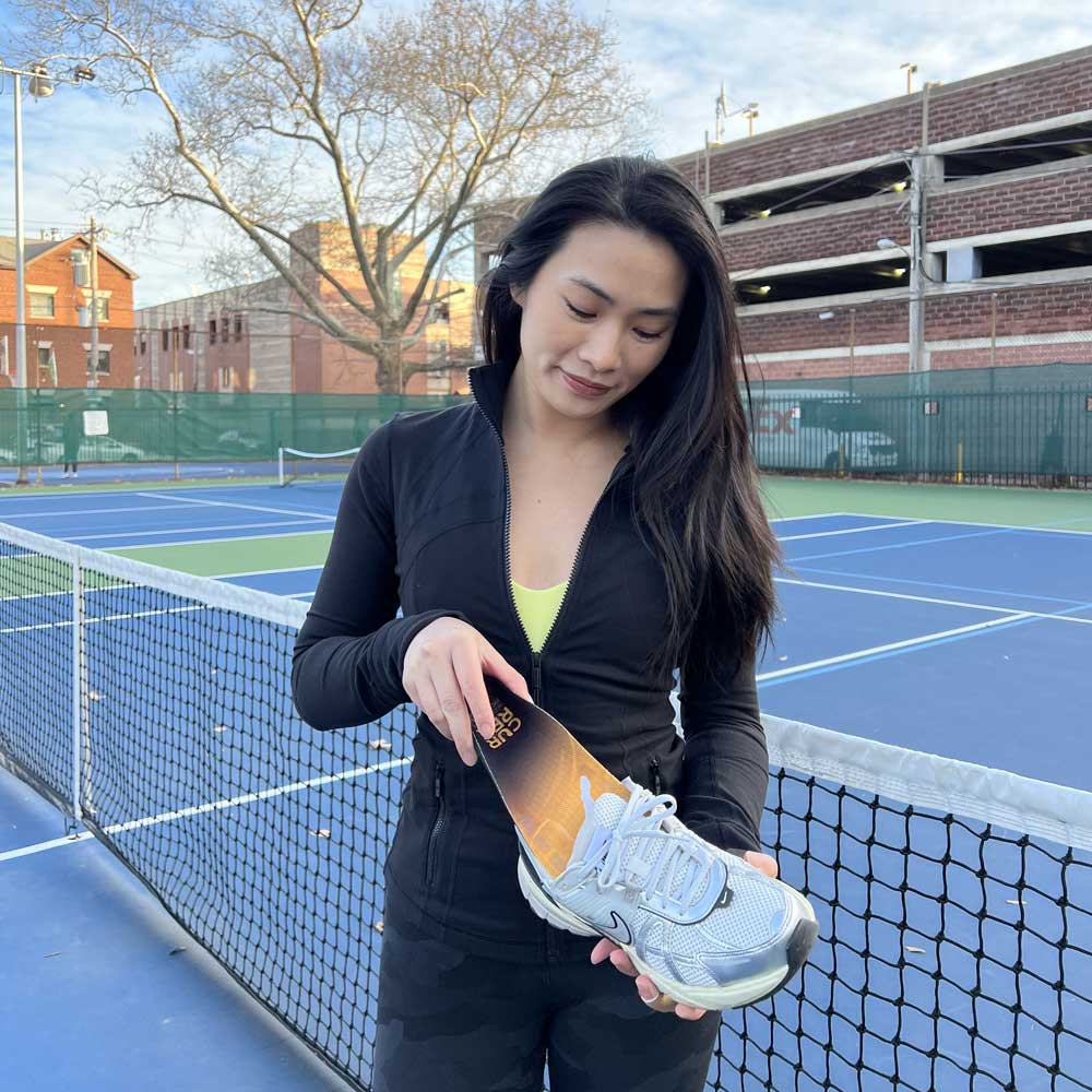 Woman on pickleball court, placing medium profile CURREX PICKLEBALLPRO insoles into white tennis shoes #profile_low
