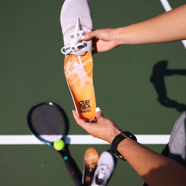 Person standing on green tennis court while inserting CURREX ACEPRO yellow medium profile insoles into gray tennis shoe #profile_medium