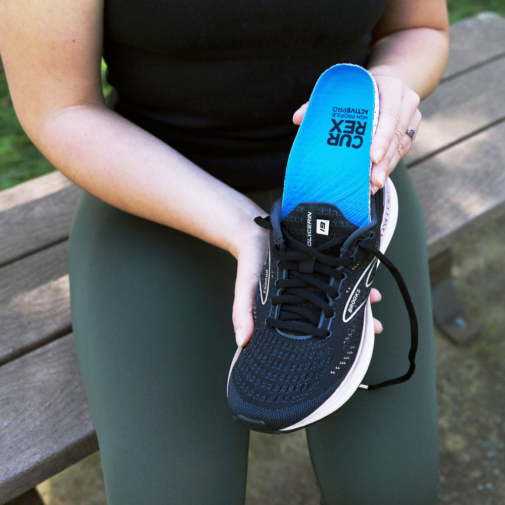 Woman sitting on a bench at a park, placing blue high profile CURREX ACTIVEPRO insoles into black sneaker #profile_high