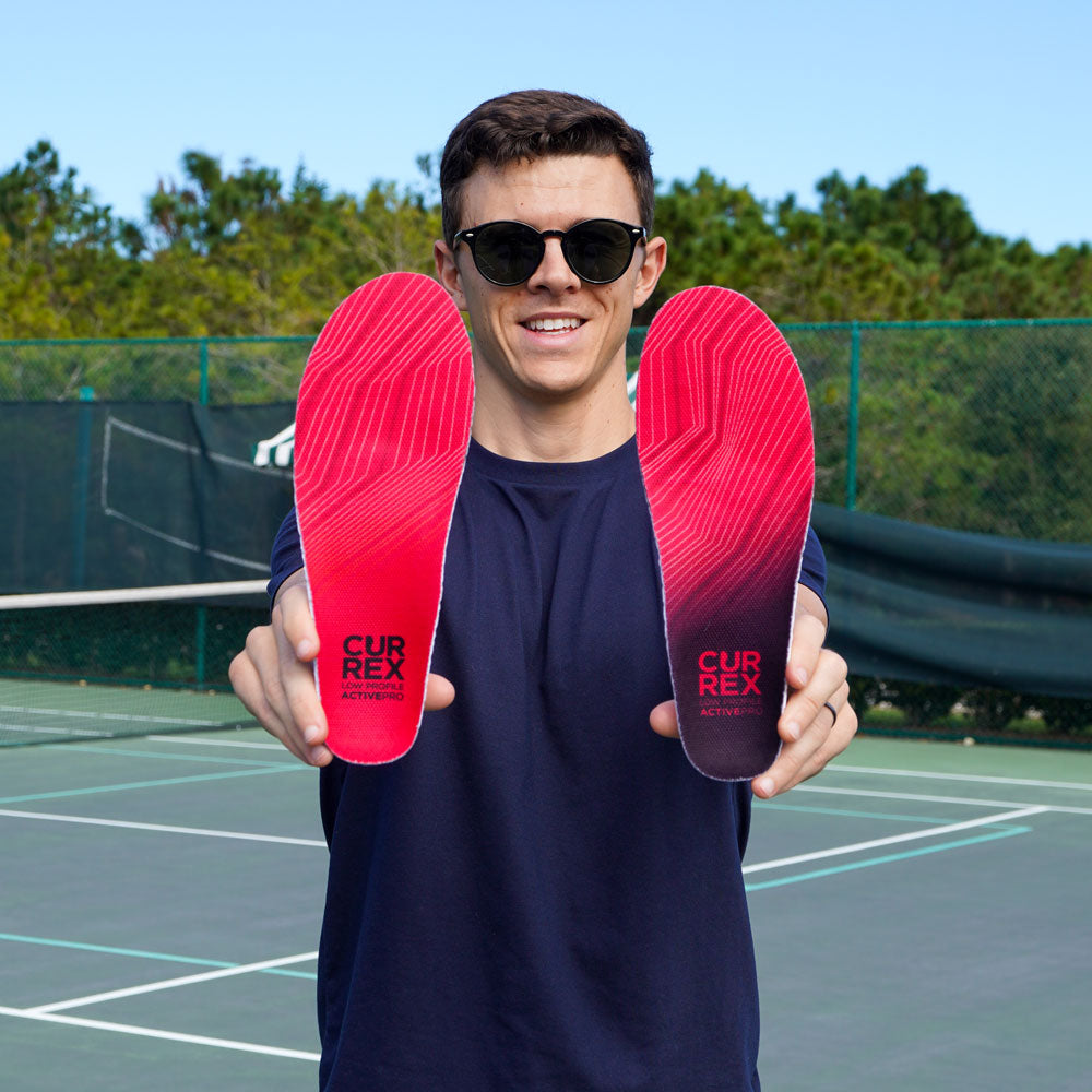 Man standing on tennis court while holding up a pair of red low profile CURREX ACTIVEPRO insoles #profile_low