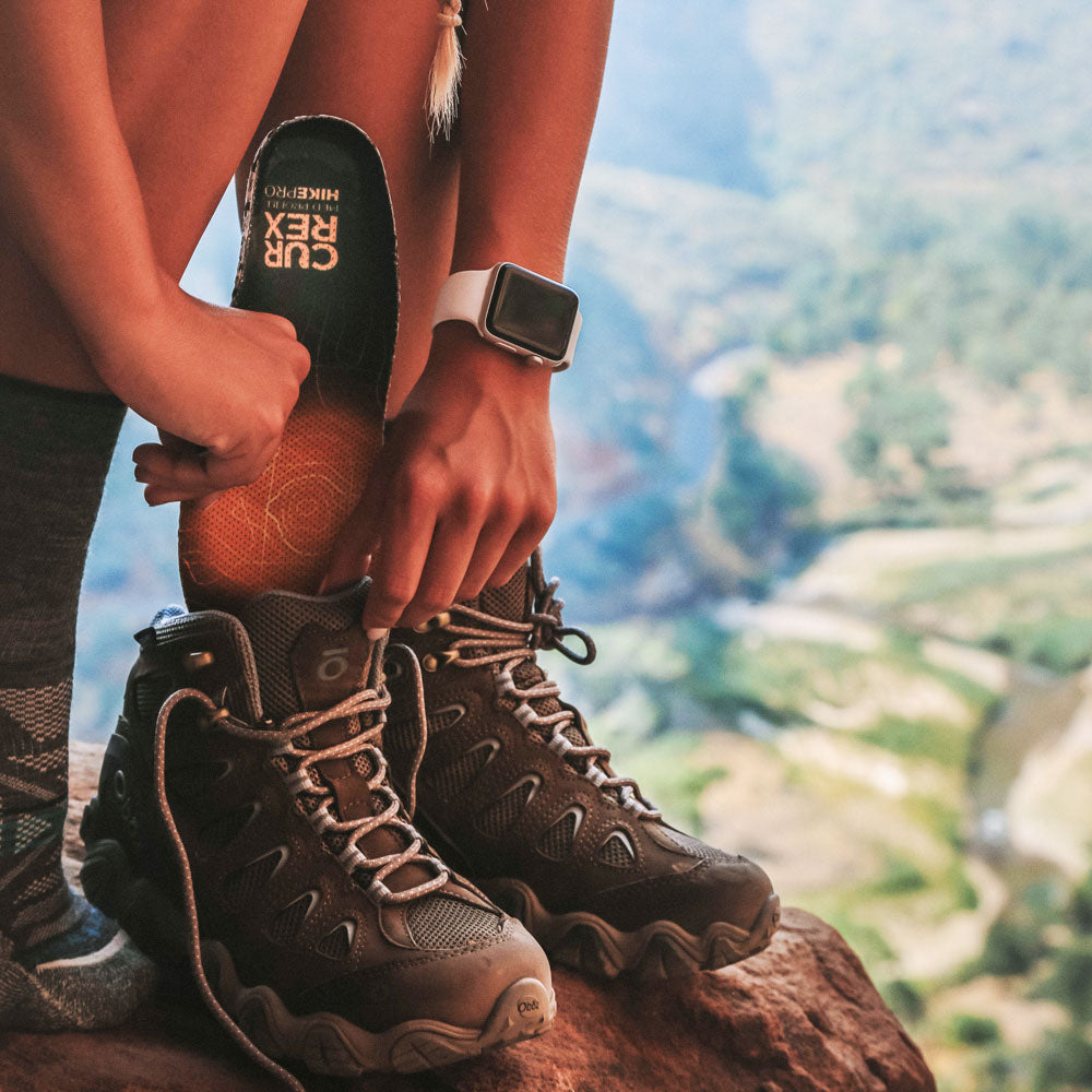 Woman placing yellow medium profile CURREX HIKEPRO insoles into hiking shoes while standing near mountain cliff #profile_medium