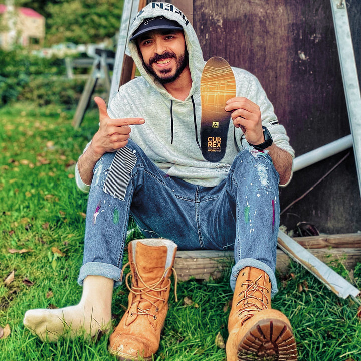 Man sitting in yard holding up a pair of CURREX WORK yellow medium profile insoles for his work boots #profile_medium